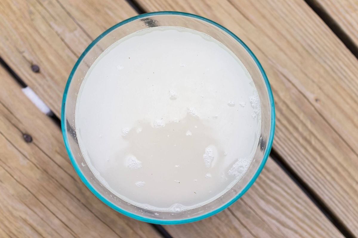 Closeup flat top lay view down of soaked rice, grain, cloudy liquid water in glass bowl on wooden background