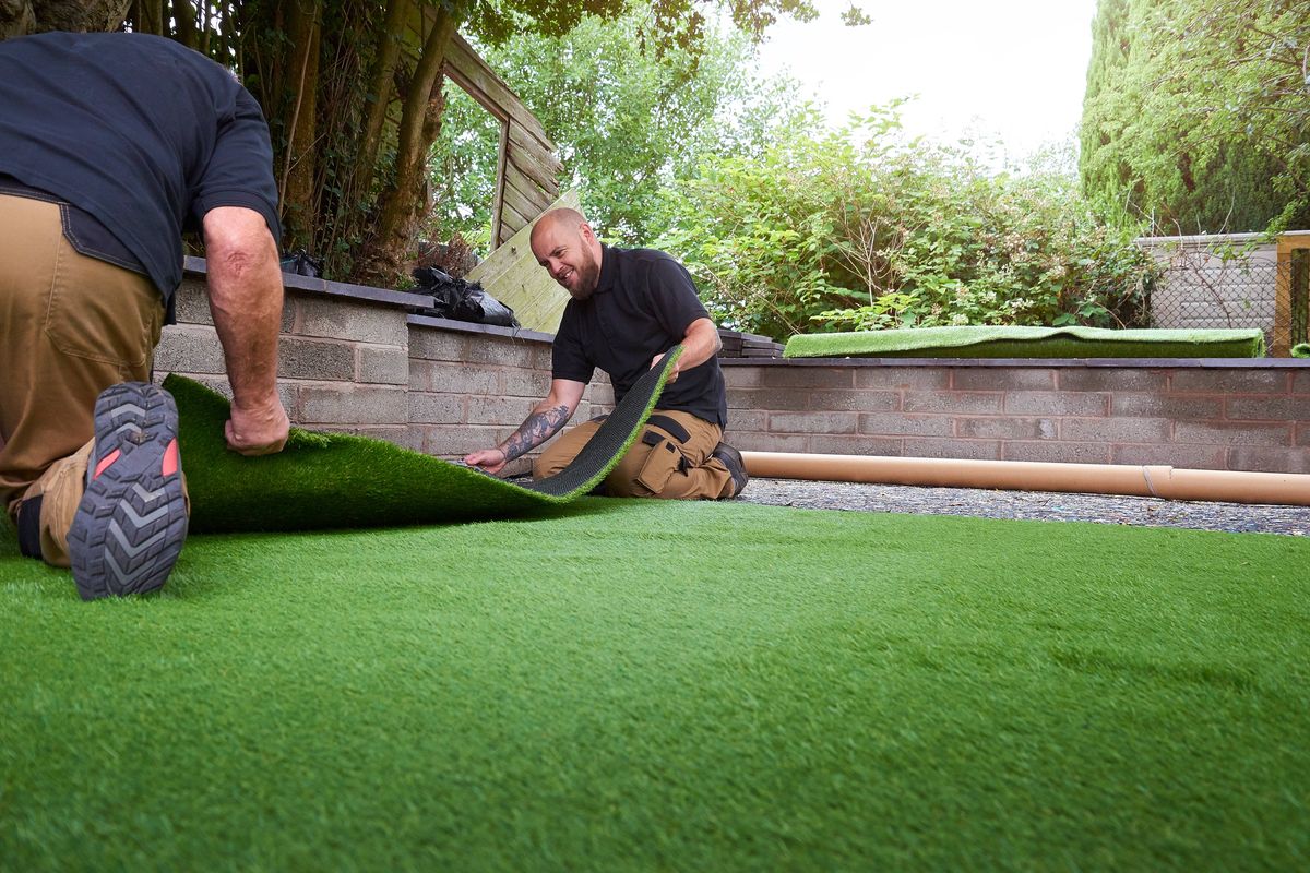 Two individuals are engaged in the process of installing artificial turf on a residential property. They are kneeling on the ground, carefully laying down the turf to cover an area that appears to be a backyard. The setting features a brick wall and lush greenery in the background, suggesting a well-maintained outdoor space.