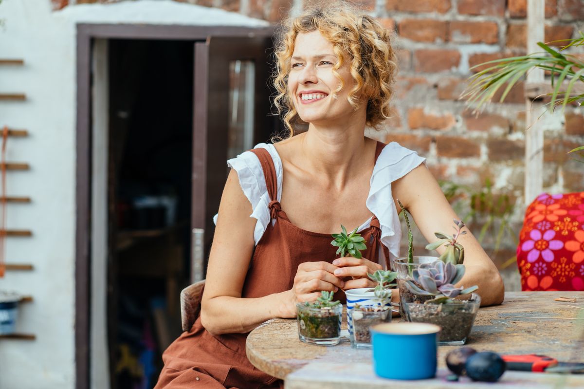 Blond haired woman gardener in brown overalls planting succulents outdoor in cosy terrace