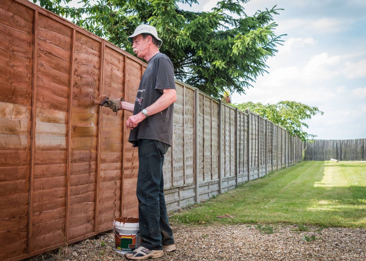 An individual is engaged in the act of applying paint to a wooden fence. The fence is situated amidst a grassy area, with a backdrop of trees and a clear sky.