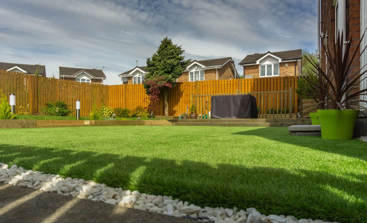 An outdoor scene depicts a well-maintained backyard featuring a lush green lawn, bordered by a stone pathway. Behind the lawn, a wooden fence extends across the background, with a large green plant in a pot to the right. Beyond the fence, a row of residential houses is visible, and the sky above is partly cloudy.