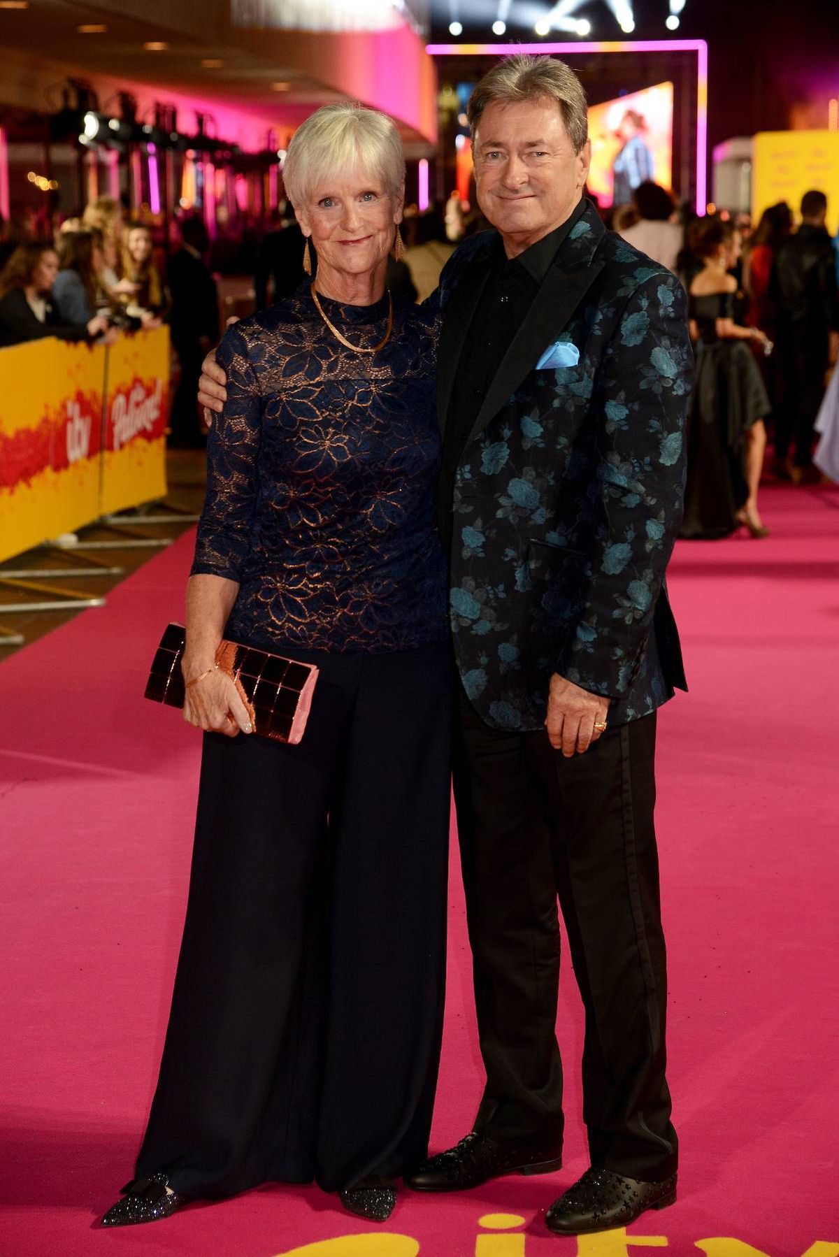 An older couple stands close together on a red carpet at an event, with a crowd of people and a vibrantly lit background behind them.