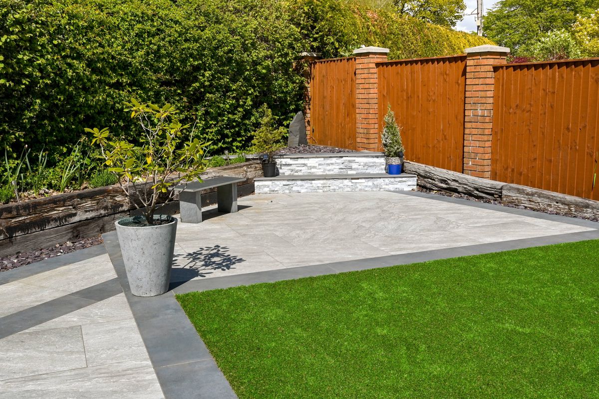 Residential back garden landscaped with light and dark grey porcelain paving slabs and artificial grass. No people.