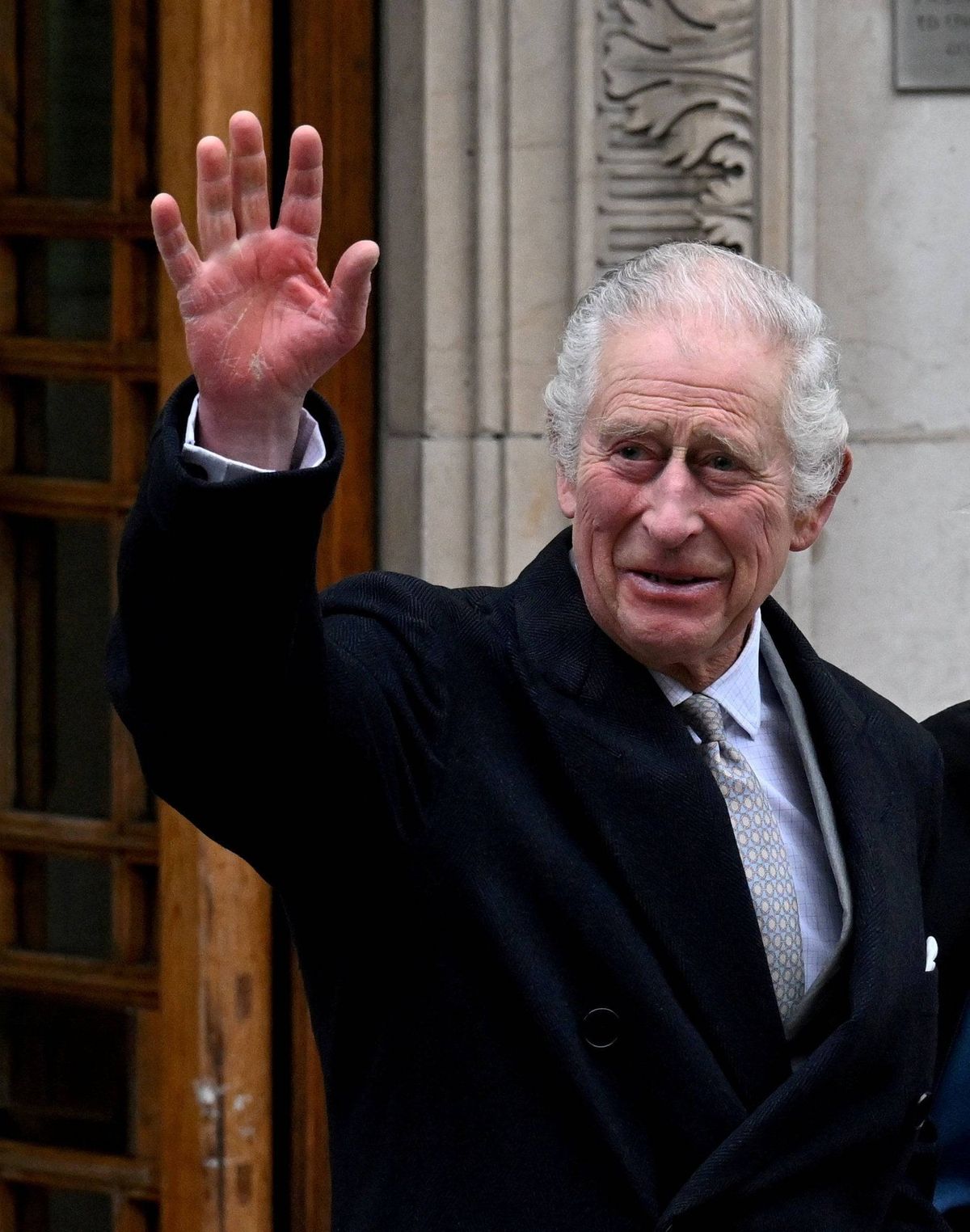An elderly gentleman dressed in formal attire is standing outside, waving his hand enthusiastically. The background features an architectural structure with ornate designs.