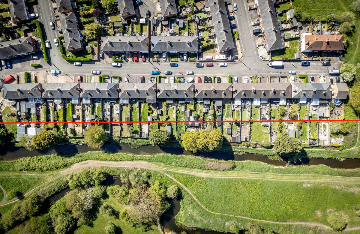 Aerial view of an urban residential area featuring numerous terraced houses, a roadway with parked vehicles, and a green landscape with trees and grass. A red line highlights a specific area within the property.