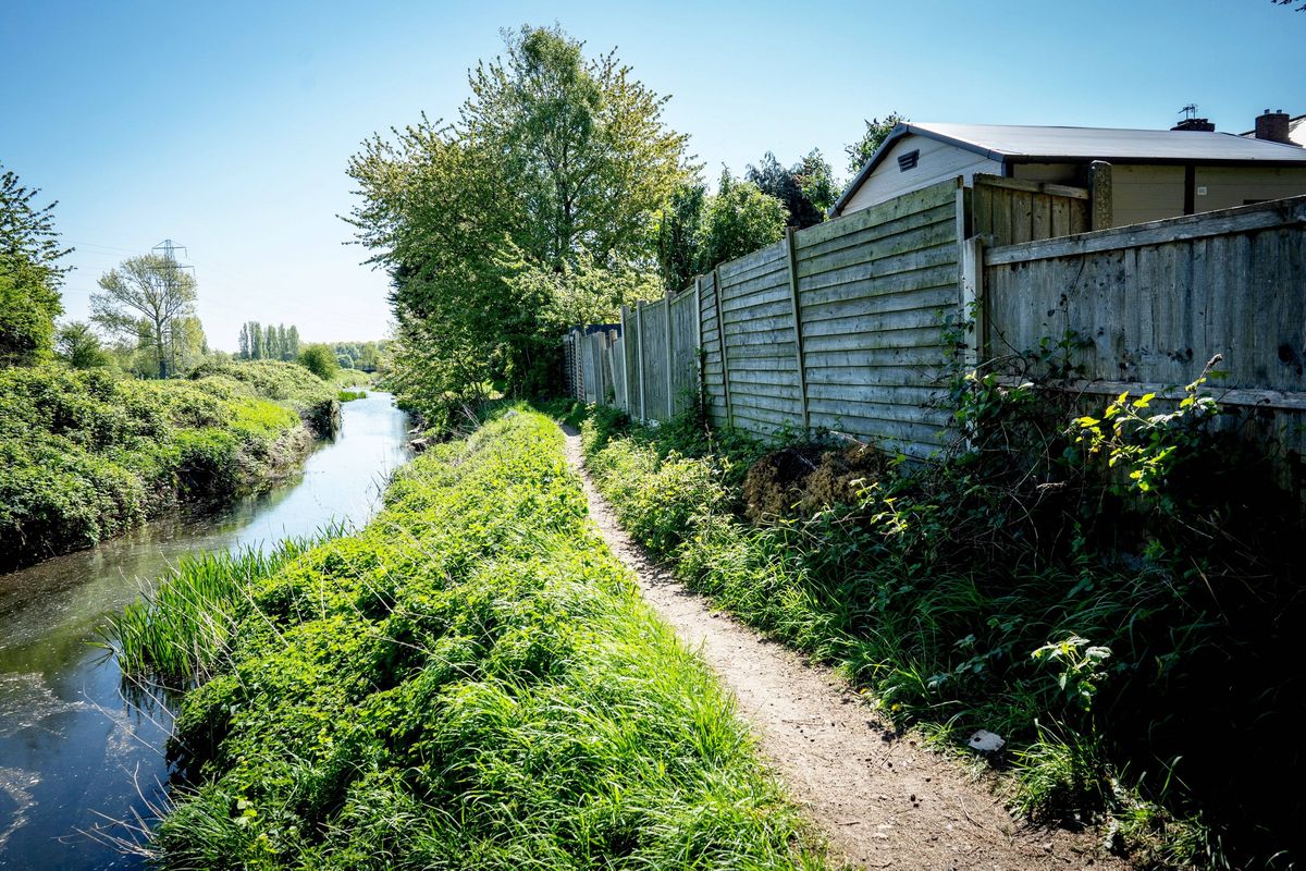 A serene rural scene with a small river flanked by lush greenery. A narrow path runs along the riverbank, bordered by a wooden fence on one side. Behind the fence, a house with a sloping roof is visible amidst a backdrop of trees. The sky is clear and blue, indicating a sunny day.