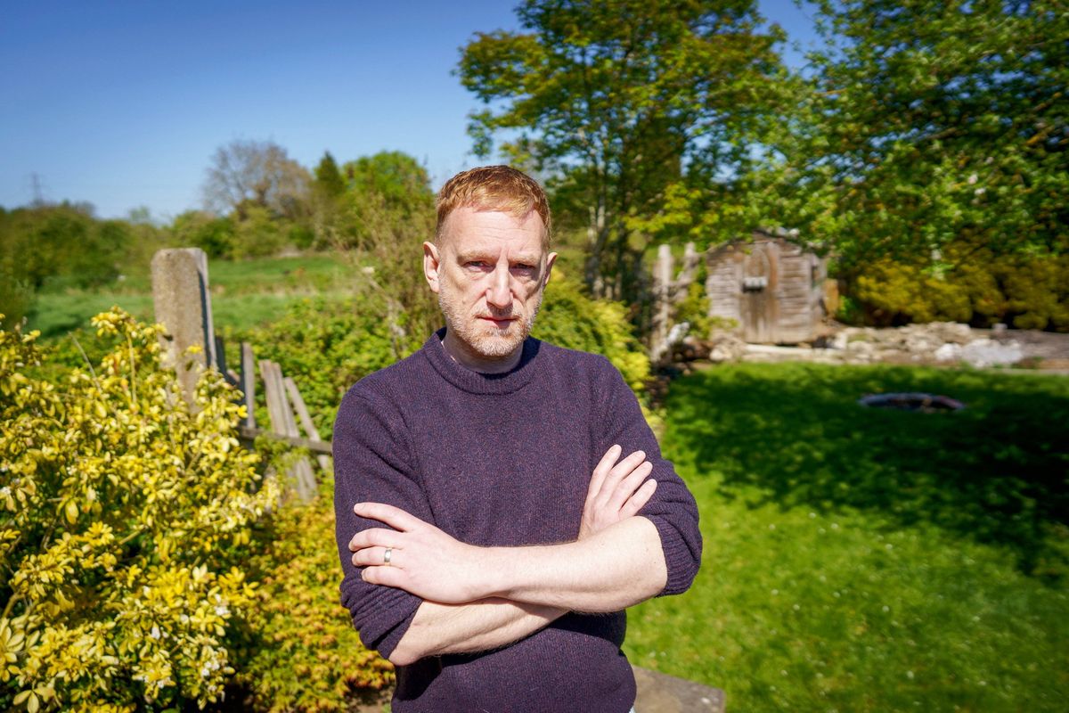 An individual with folded arms standing in a green landscape with a wooden fence and yellow flowers in the foreground.
