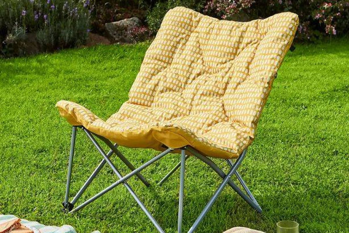 An outdoor folding chair with a yellow cushion is positioned on a lush, green lawn, accompanied by various plants in the background.