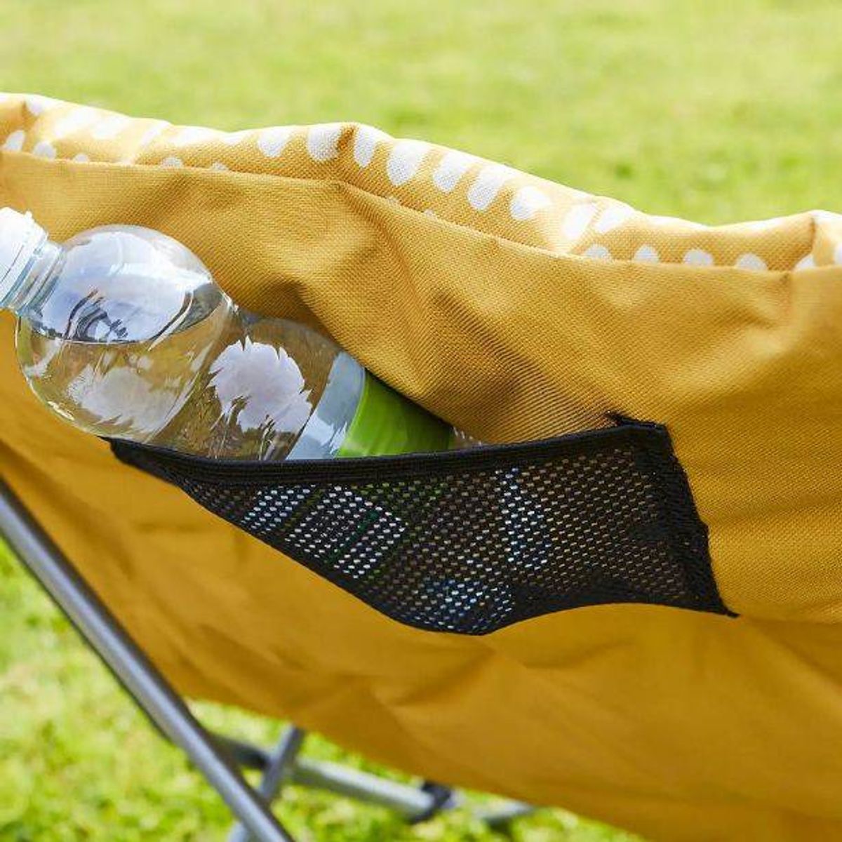 A close-up view of a yellow folding chair with a mesh pocket, containing a bottle of water and a transparent container, placed on a grassy surface.