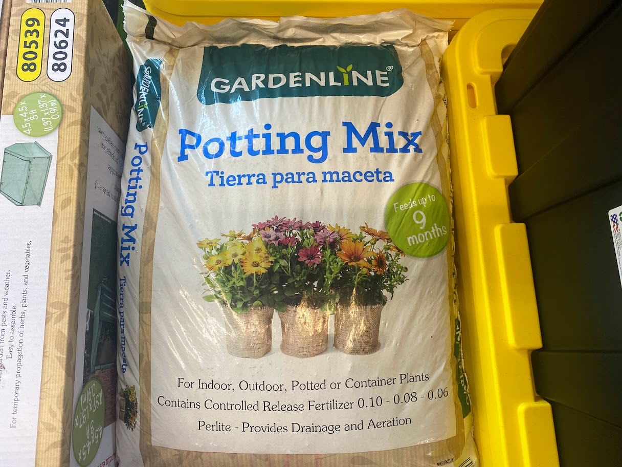 A bag of Gardenline Potting Mix with images of potted flowers on the front, labeled for indoor, outdoor, potted, or container plants. The yellow edge of a storage bin is visible on the right.