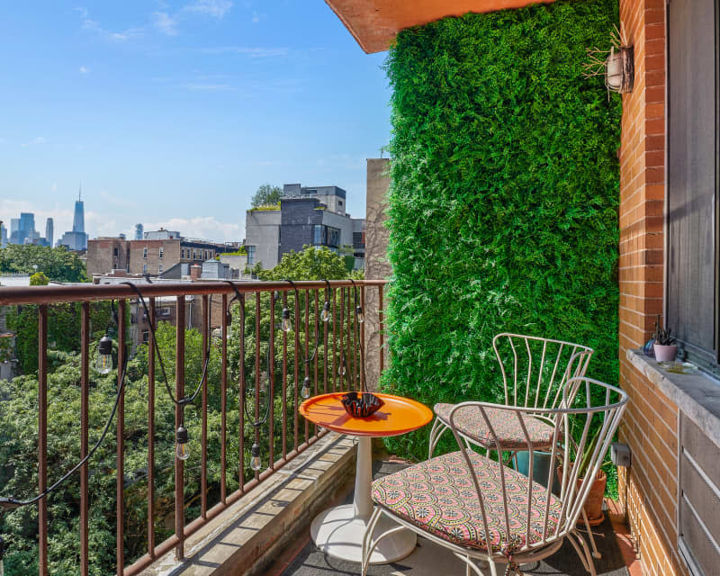 City balcony with orange table, patterned chair, green wall, and view of skyline and trees.