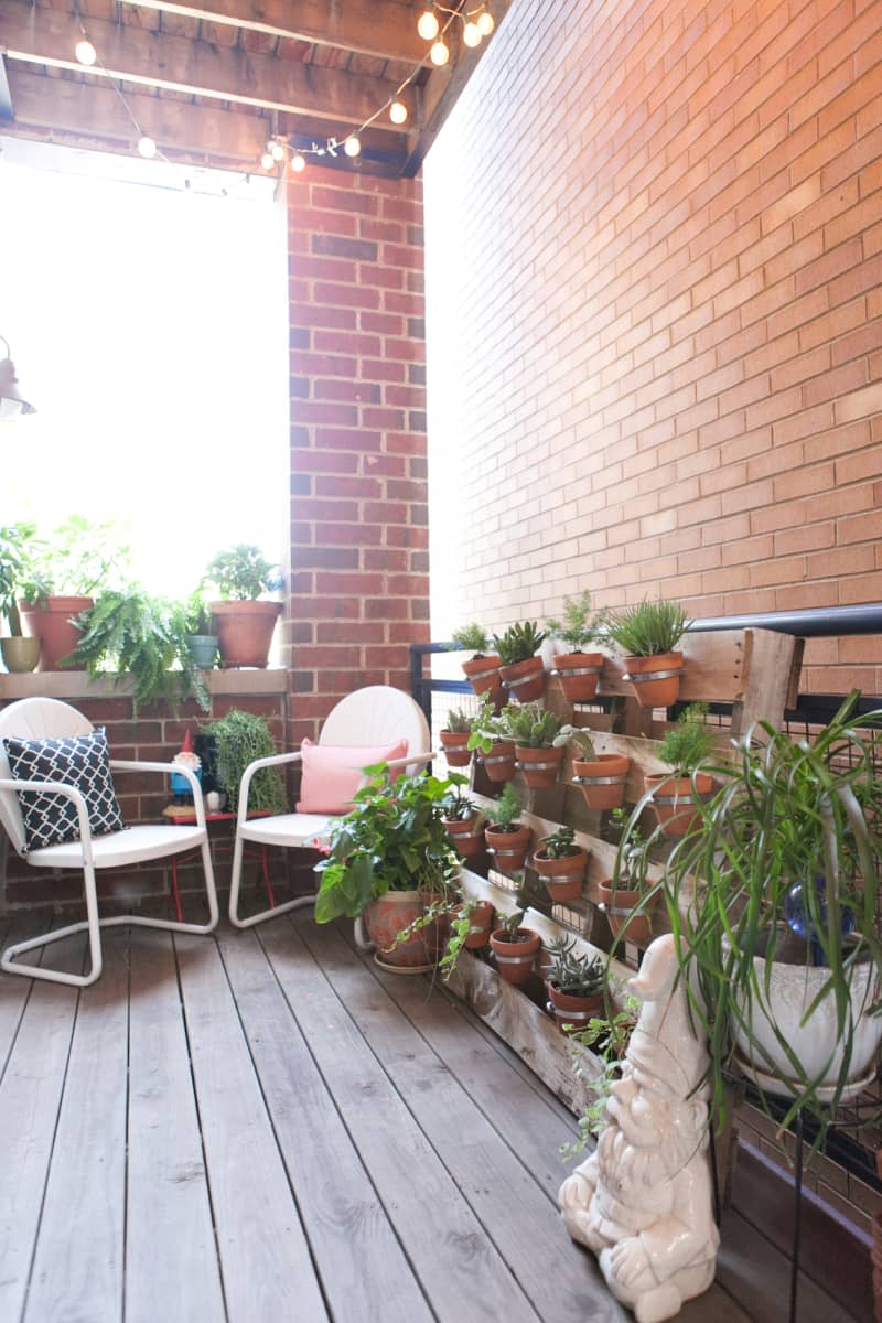 Cozy balcony with potted plants, string lights, white chairs, and a garden gnome statue.