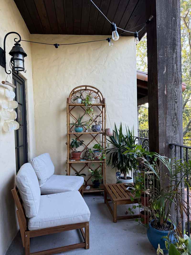 Cozy balcony with two light-colored chairs, a wooden table, and a plant shelf filled with various potted plants.