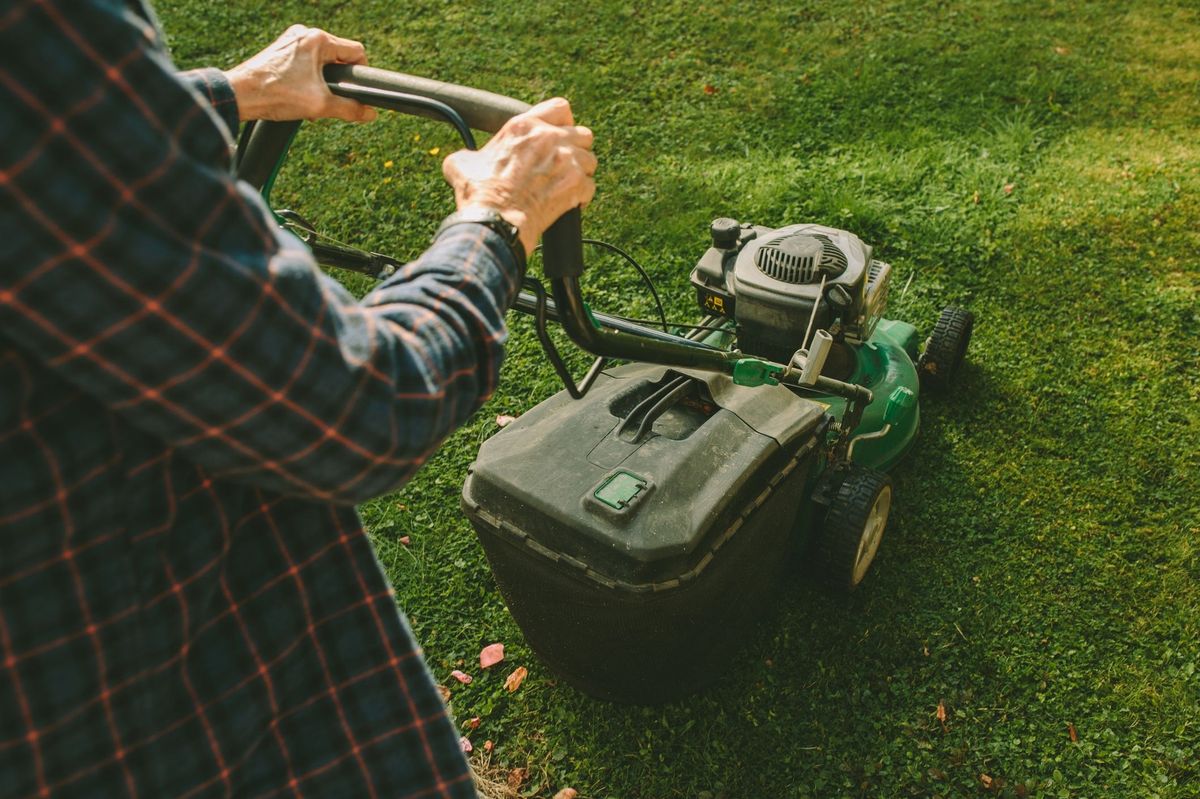 A senior woman operates a push lawn mower in a rustic backyard