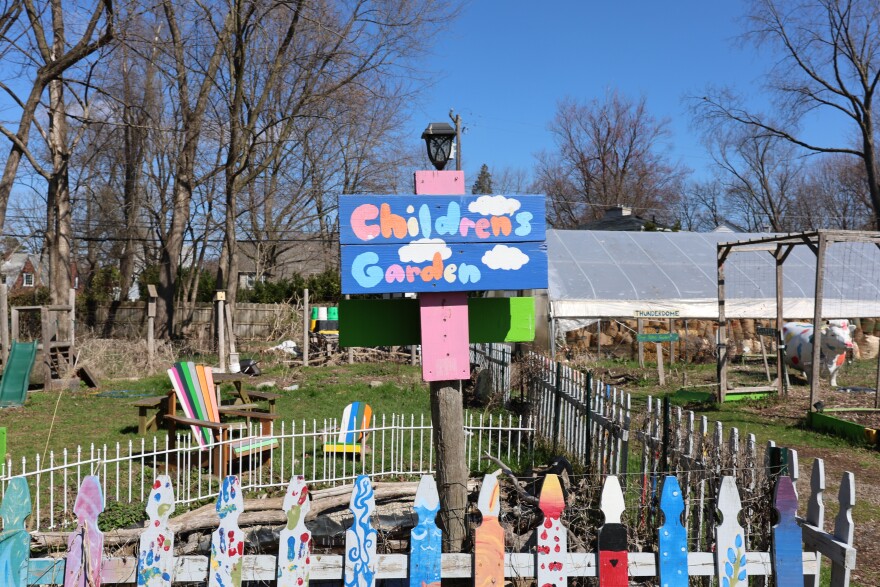 A colorful blue, pink and orange hand-painted sign on a post reads "Children's Garden." A hand-painted fence circles a plot of dirt, with a hoop house visible in the background.