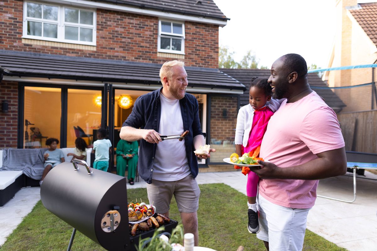 Two individuals are engaged in conversation while preparing food in an outdoor backyard setting. A grill and a house are visible in the background.