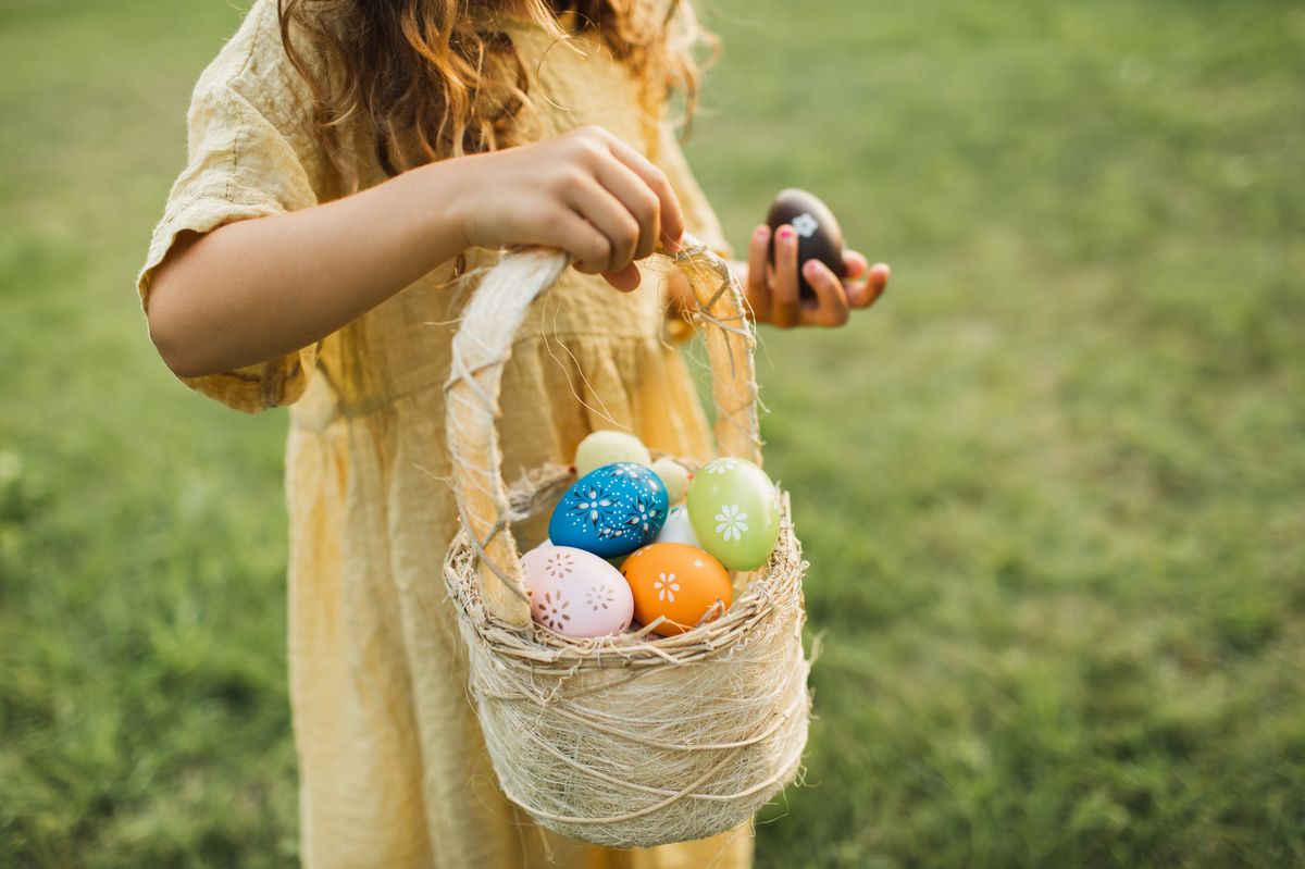 Cute little girl with bunny ears holding an easter egg basket and finding eggs in the grass on green lawn. Little girl wearing fluffy Bunny ears 
gathering colorful egg in park in wicker basket.  Easter concept.