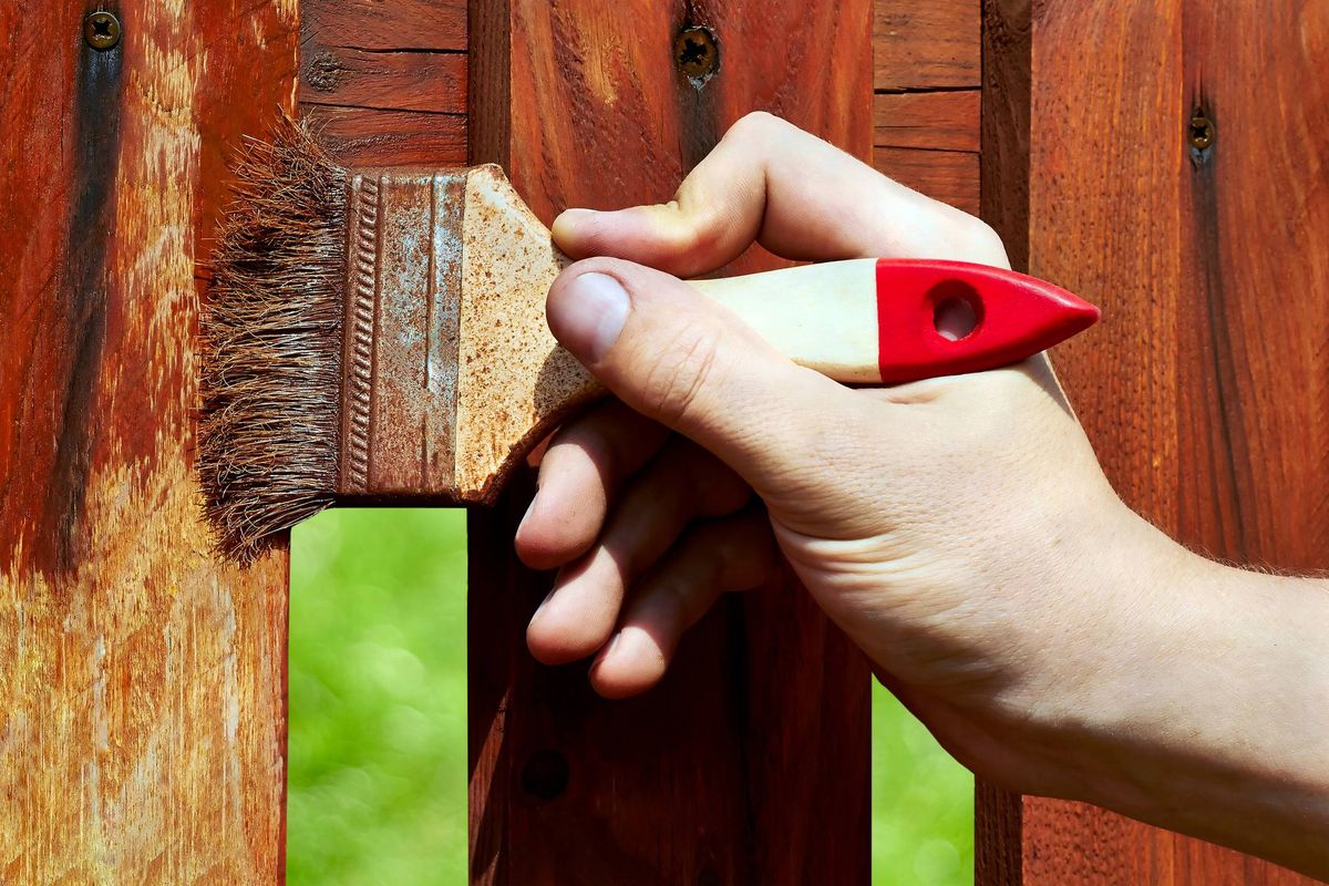A hand is holding a red and white paintbrush, applying paint to a wooden fence in an outdoor setting.