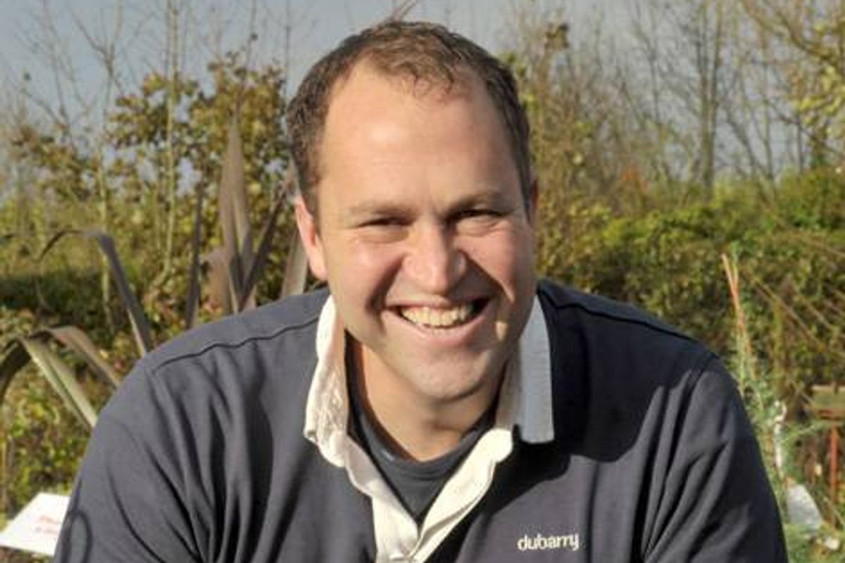 A man wearing a dark blue polo shirt with a white collar is standing in an outdoor setting, surrounded by greenery, and is smiling broadly at the camera.