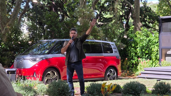 Jamie Durie holding a microphone, one hand in the air and standing in a garden. There's a red van in the background.
