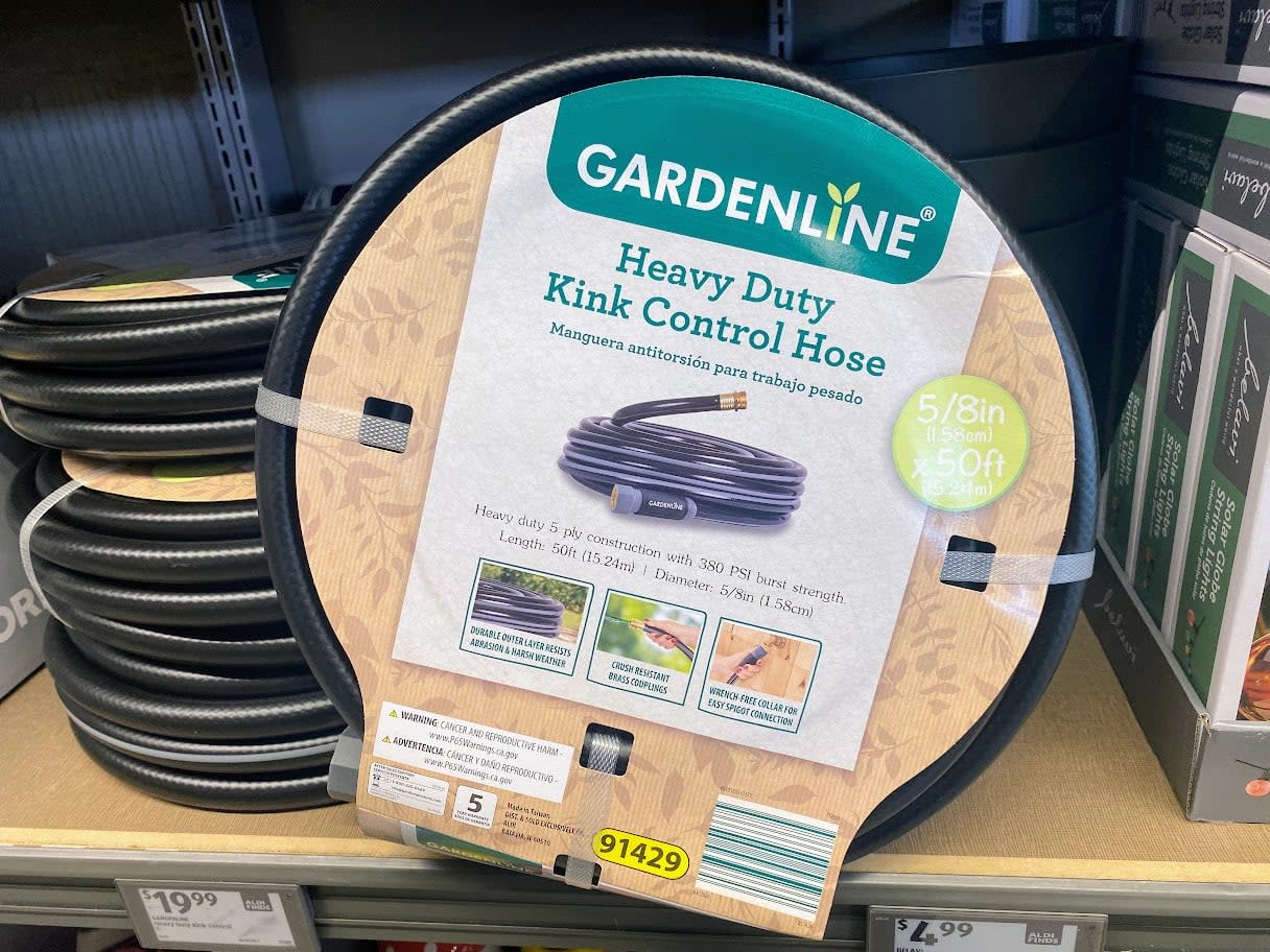 A coiled Gardenline Heavy Duty Kink Control Hose in packaging is displayed on a store shelf. The label highlights it is 5/8 inch wide and 50 feet long, showing features and product information.