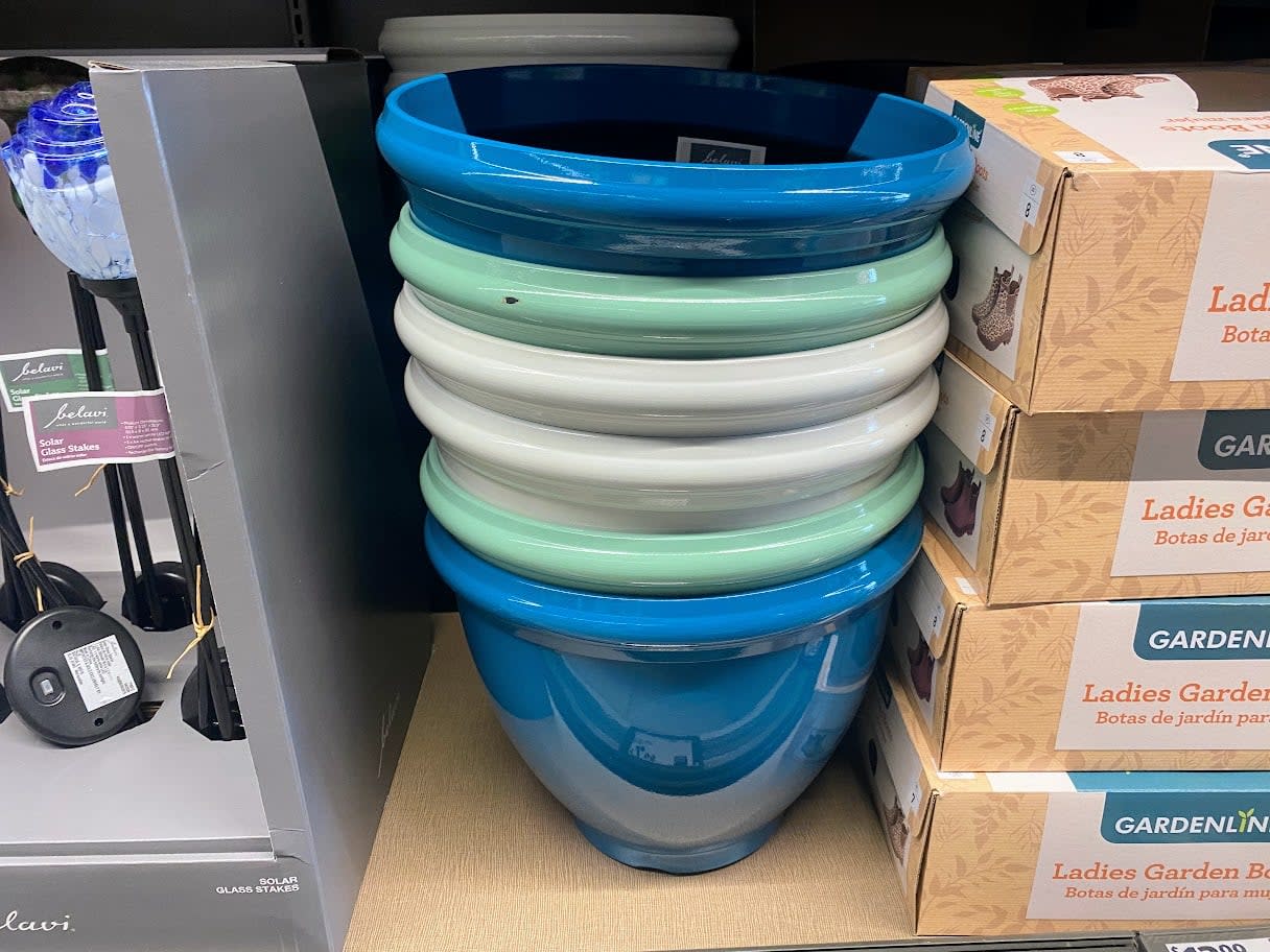 A stack of colorful ceramic plant pots, primarily blue, green, and white, displayed on a store shelf next to boxed garden clogs and other gardening items.
