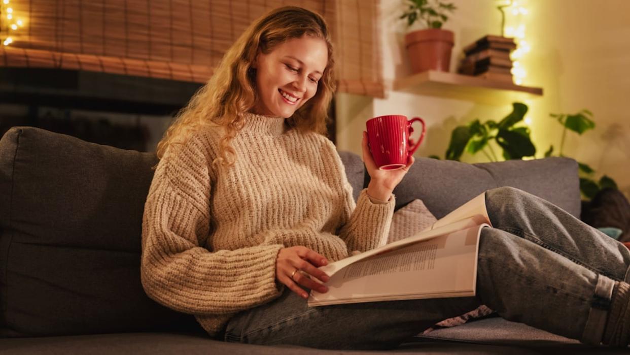 Young pretty curly young blonde woman lying on sofa at home reading magazine, cozy Christmas holidays