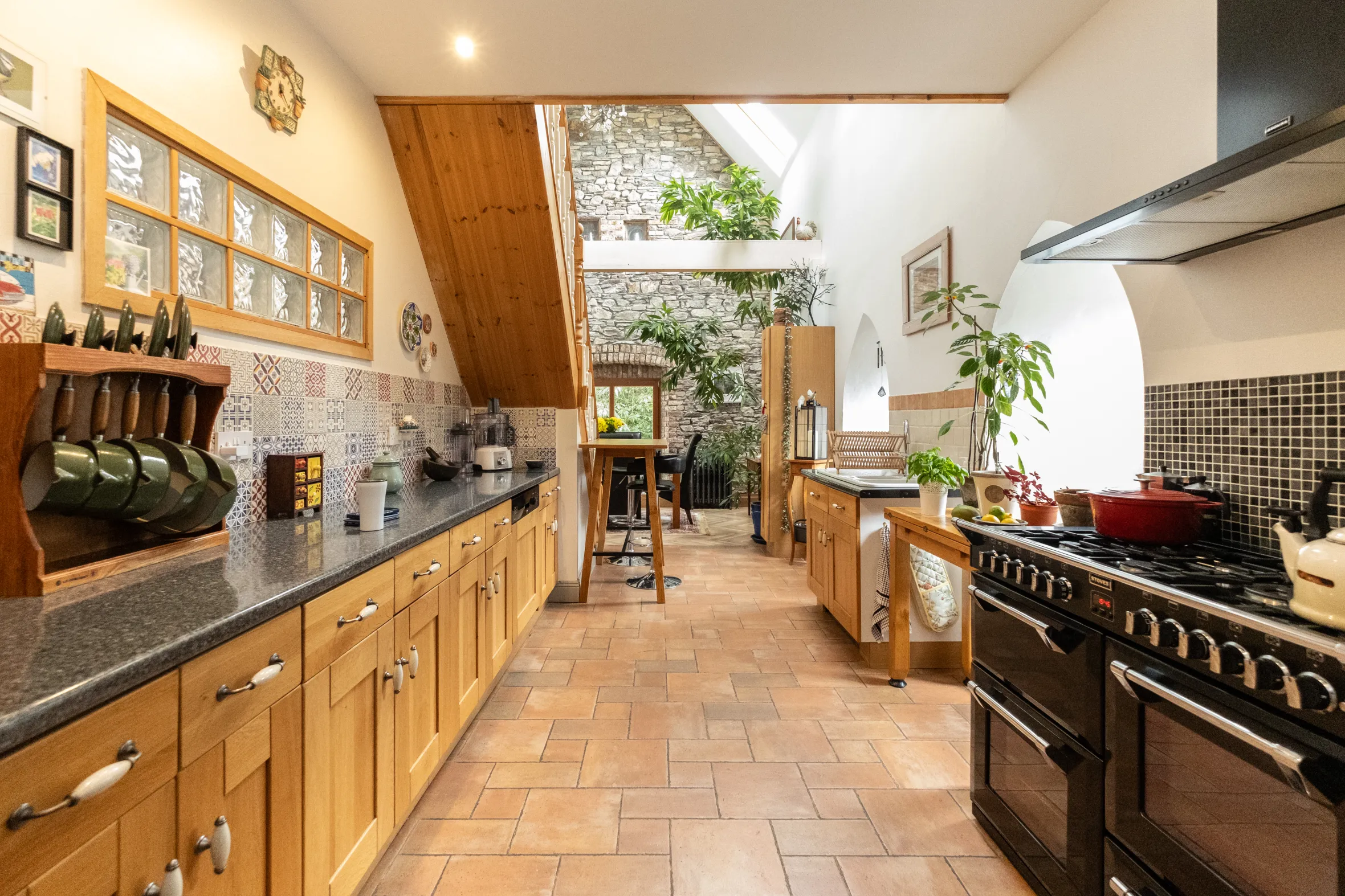 A long, brightly lit kitchen with light wood cabinets, dark countertops, and a patterned tile backsplash, leading to an open dining area.
