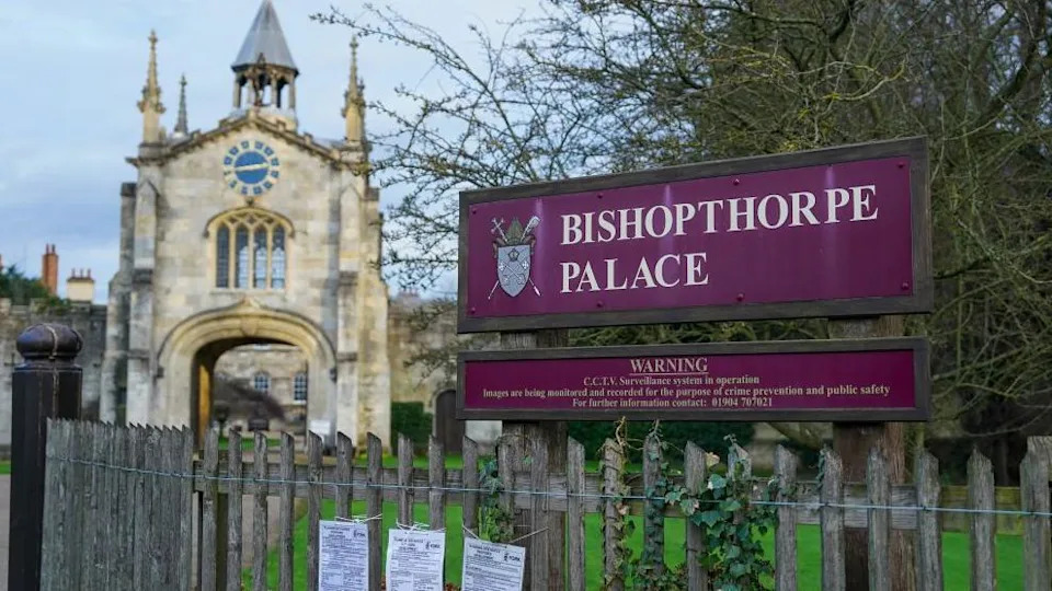 Entrance gate to to Bishopthorpe Palace, the home of the Archbishop of York, includes a stone gatehouse with a clock in the distance with a purple sign stating the name of the building and the coat of arms of the Archbishop