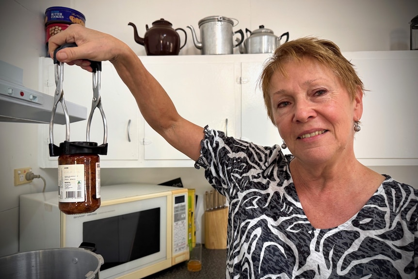 A smiling, middle-aged woman with short fair hair uses a utensil to hold up a jar while standing in a kitchen.