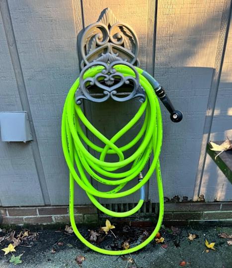 A green garden hose coiled around a decorative wall-mounted holder with fallen leaves on the ground