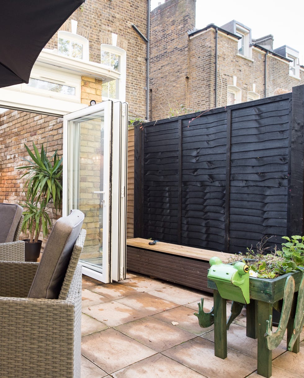 Black fence in small garden a view looking from the back garden into the dining room lounge through open bi fold patio doors with frog styled planter, paving slab patio, black timber fence, square box style rattan table and chairs with parasol