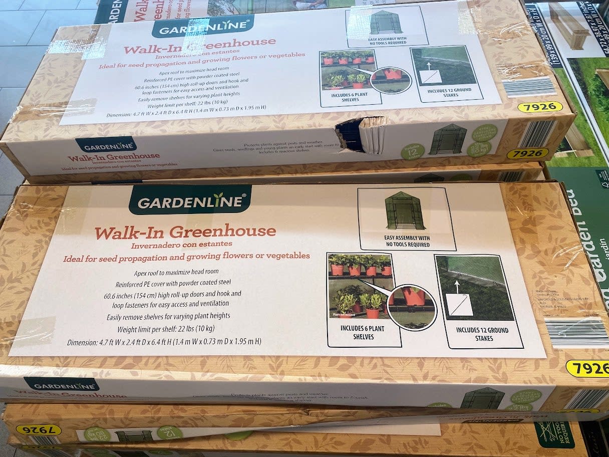 Boxes of Gardenline Walk-In Greenhouse kits are stacked on display. The packaging shows images of the assembled greenhouse, plant shelves, and ground stakes, with product details and yellow price tags labeled 7926.