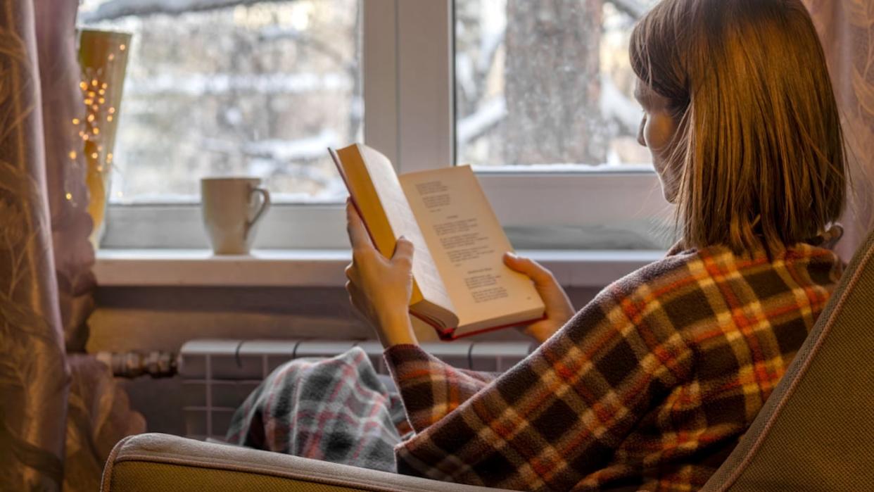 Woman with brown hair is sitting in soft armchair, wrapped in plaid, and reading book. Outside window are winter trees in snow. Cozy solitude. Back view.