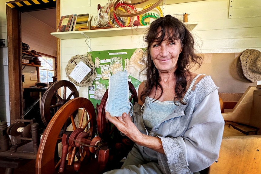 A smiling, dark-haired woman holds up a piece of fabric while sitting next to a loom.