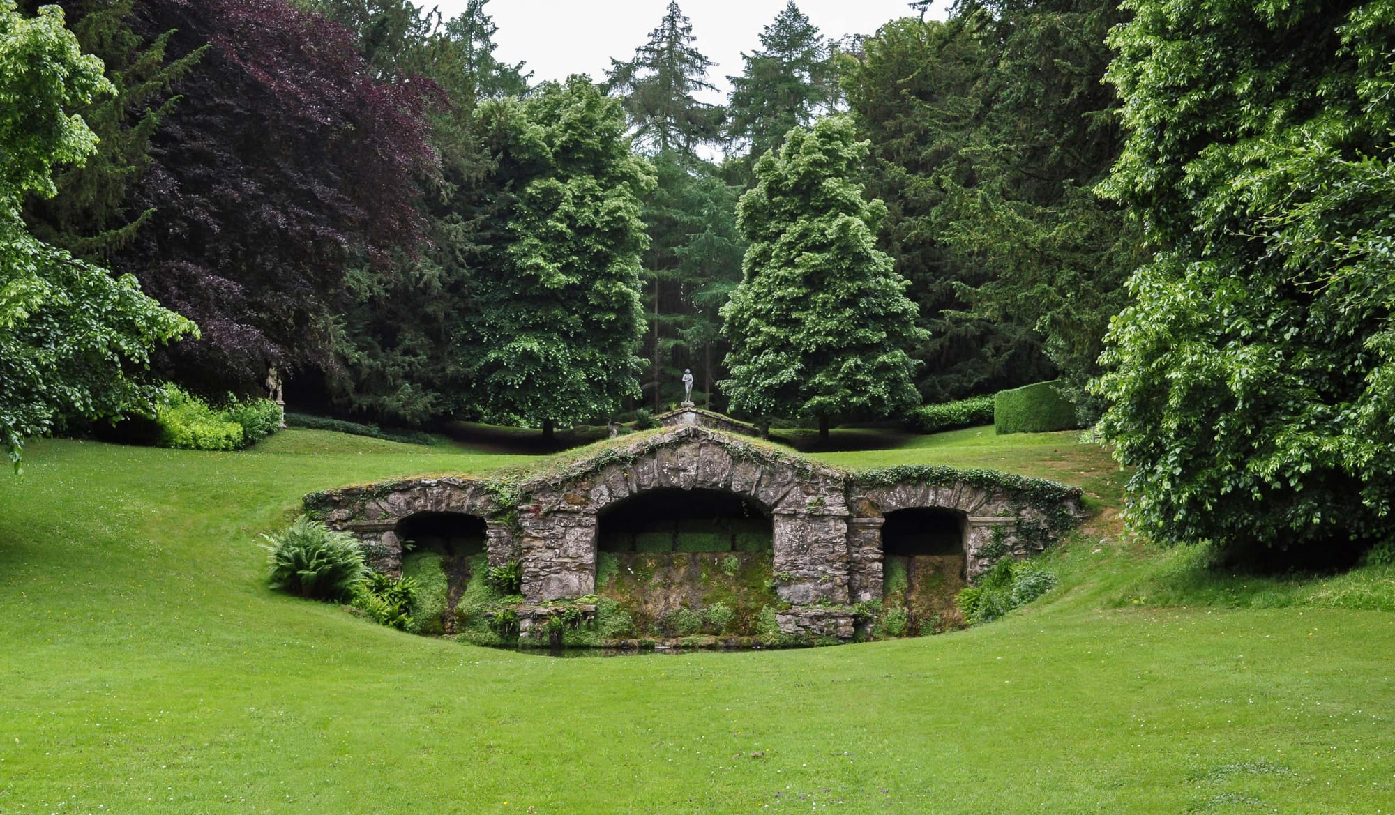 Stone garden grotto with arches at Rousham House & Garden. Lush green lawn, mossy stone architecture, and dense woodland trees in a classic English landscape garden design.