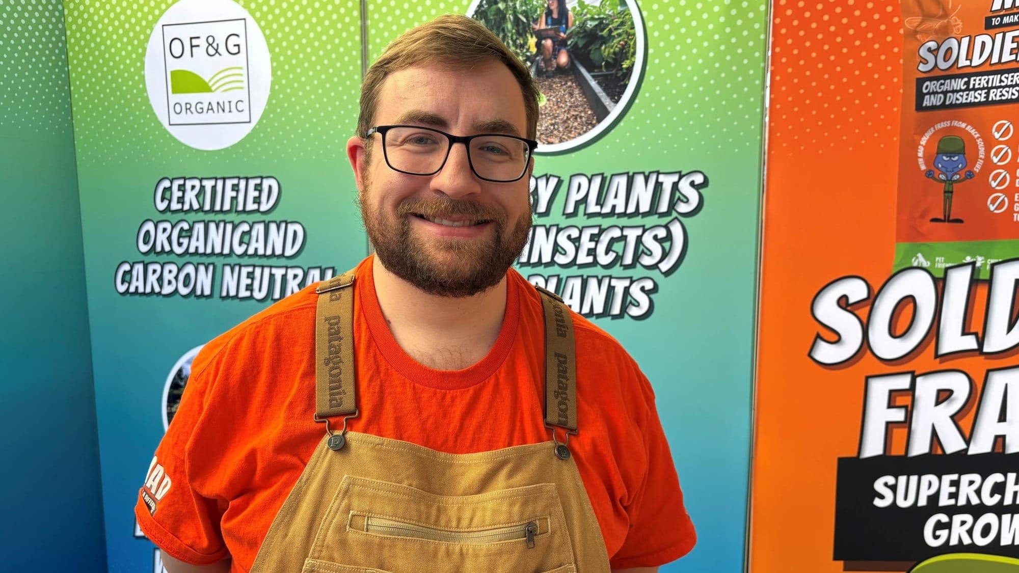 Dr David Stanford-Beale wears a bright orange t-shirt and tan dungarees, standing in front of colourful gardening displays for organic and carbon-neutral products.
