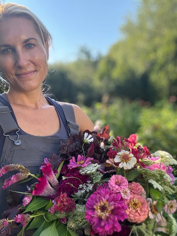 Meet the Nashville Grower Behind Bloom & Bounty - 2 Meredith Bishop stands outdoors in gray overalls, her FACE lit with a smile as she holds a large bouquet of colorful flowers; greenery and trees fill the background.