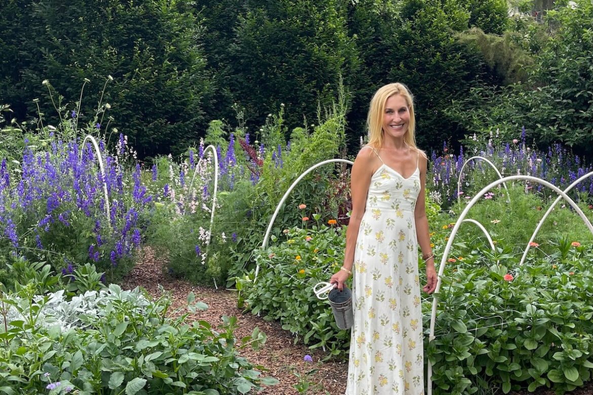 Meredith Bishop is Behind the Garden You WISH You Had Meredith Bishop, wearing a white floral dress, stands in a garden among green plants and purple flowers, her FACE serene as she holds a watering can.
