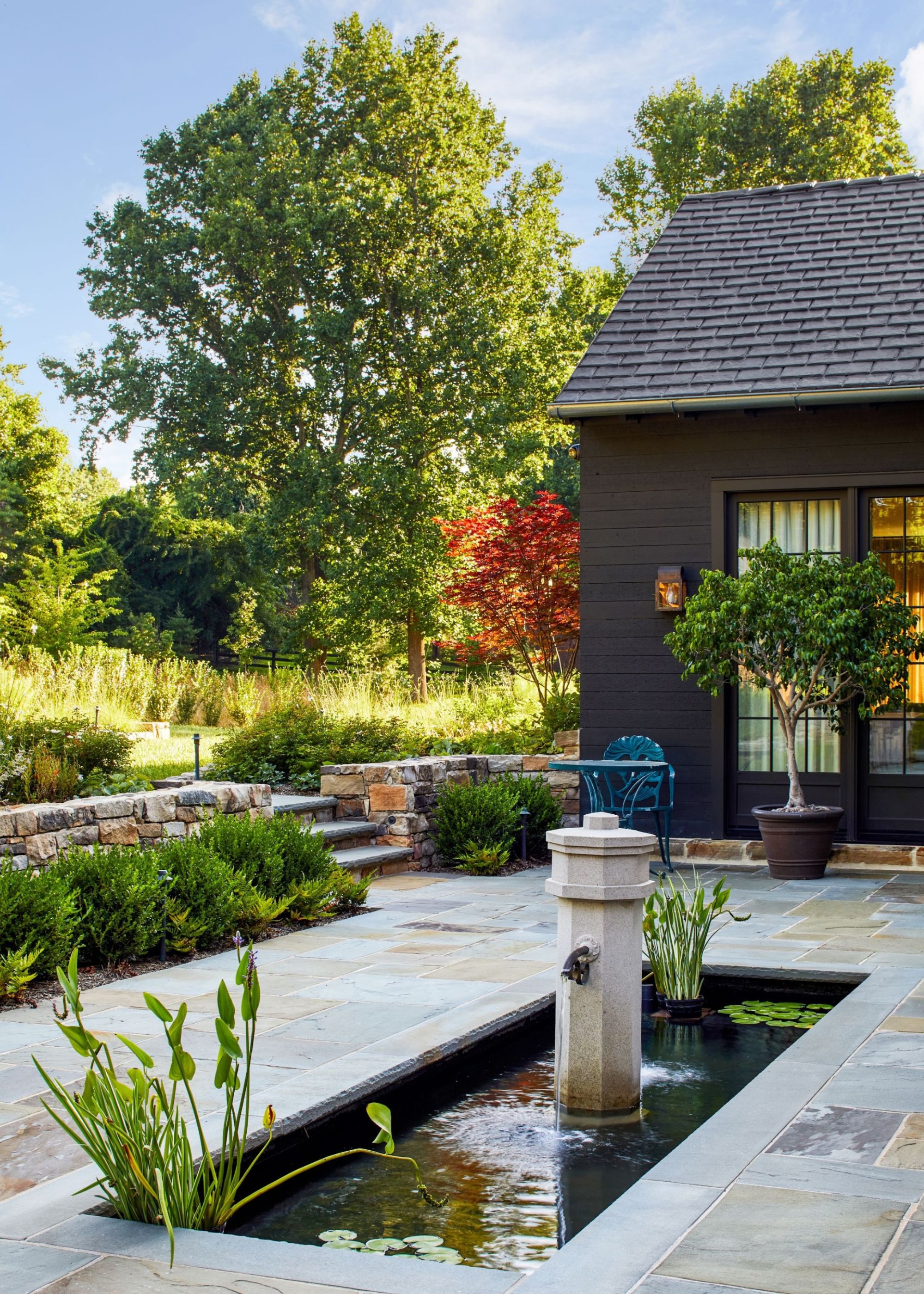 A calming garden with a central water feature built into the ground with trees and greenery in the background behind the dark-painted house