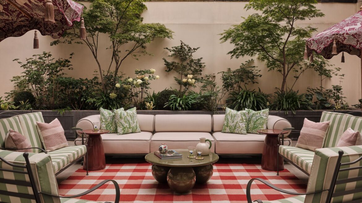 Why Designers Are ‘Upholstering’ Their Gardens In 2026 Garden with plants growing on the wall, a large red gingham rug with a large cream sofa on top, green metal armchairs with striped green cushions, and a marble green round coffee table at the centre.