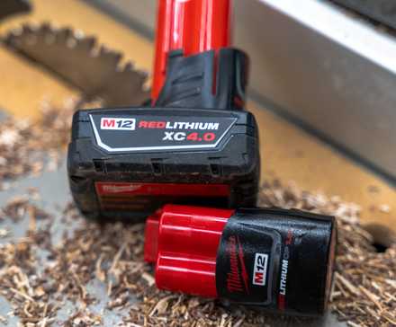 Two Milwaukee M12 batteries sitting on a table saw with sawdust and a blade in the background.