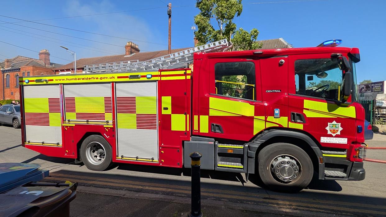 A side view of a fire engine. The red, yellow and silver vehicle is stationary on a road with a red brick building in the background. The scene is set against a blue sky and sunshine.