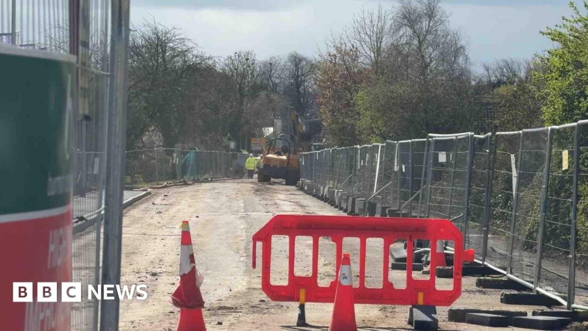 The photo is framed by metal construction fencing on either side. Down the centre we can see a road with its surface stripped away. Cones and red barriers are scattered about, and yellow construction vehicles can be seen in the distance, as well as workers in high vis clothing