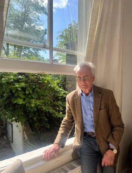 An older person standing indoors by a large window, with thick vegetation visible outside filling the neighbouring garden.
