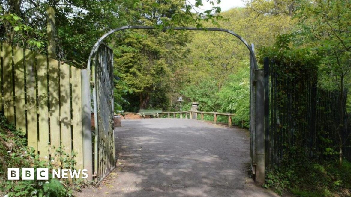 Metal gates at one of the entrances to Saltburn's Valley Gardens. They are open, leading on to a path with trees in the background. Wooden fences stand either side of the gates.