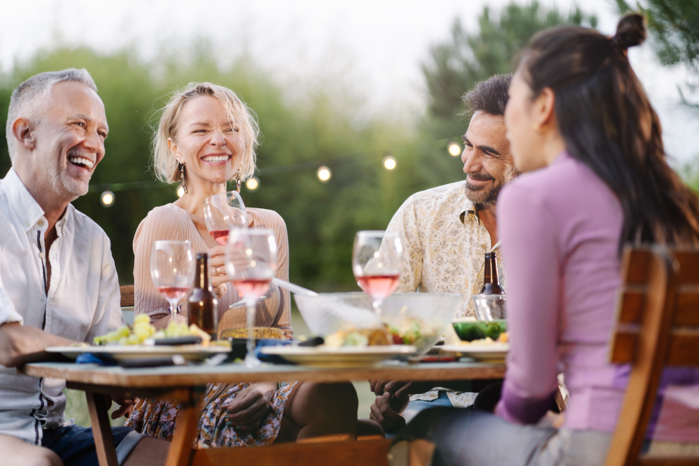 Family friends having dinner around table in a garden in summer, drinking wine, talking and laughing in outdoor restaurant at night