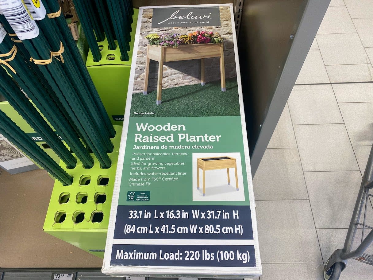 A box for a belavi wooden raised planter on a store shelf, displaying dimensions, features, and a photo of the assembled planter with plants inside. Text indicates a maximum load of 220 lbs (100 kg).