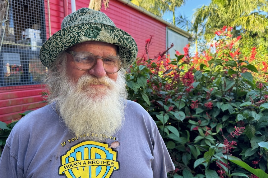 A smiling man with a long, white beard stands in a garden near a brightly-coloured shed.