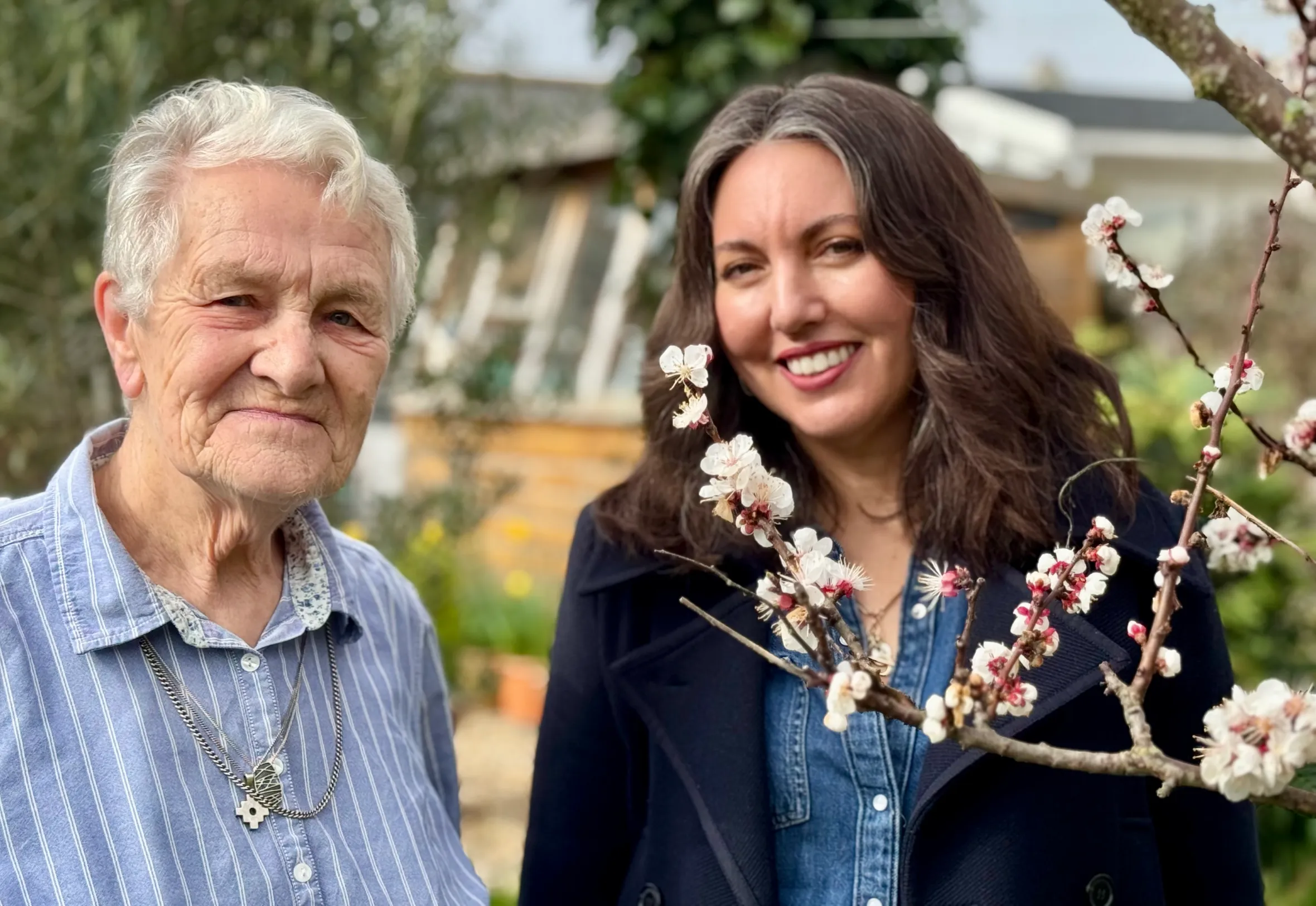 Melanie Hick and Shirley Thompson smile at the camera while standing outside with flowering tree branches in the foreground.